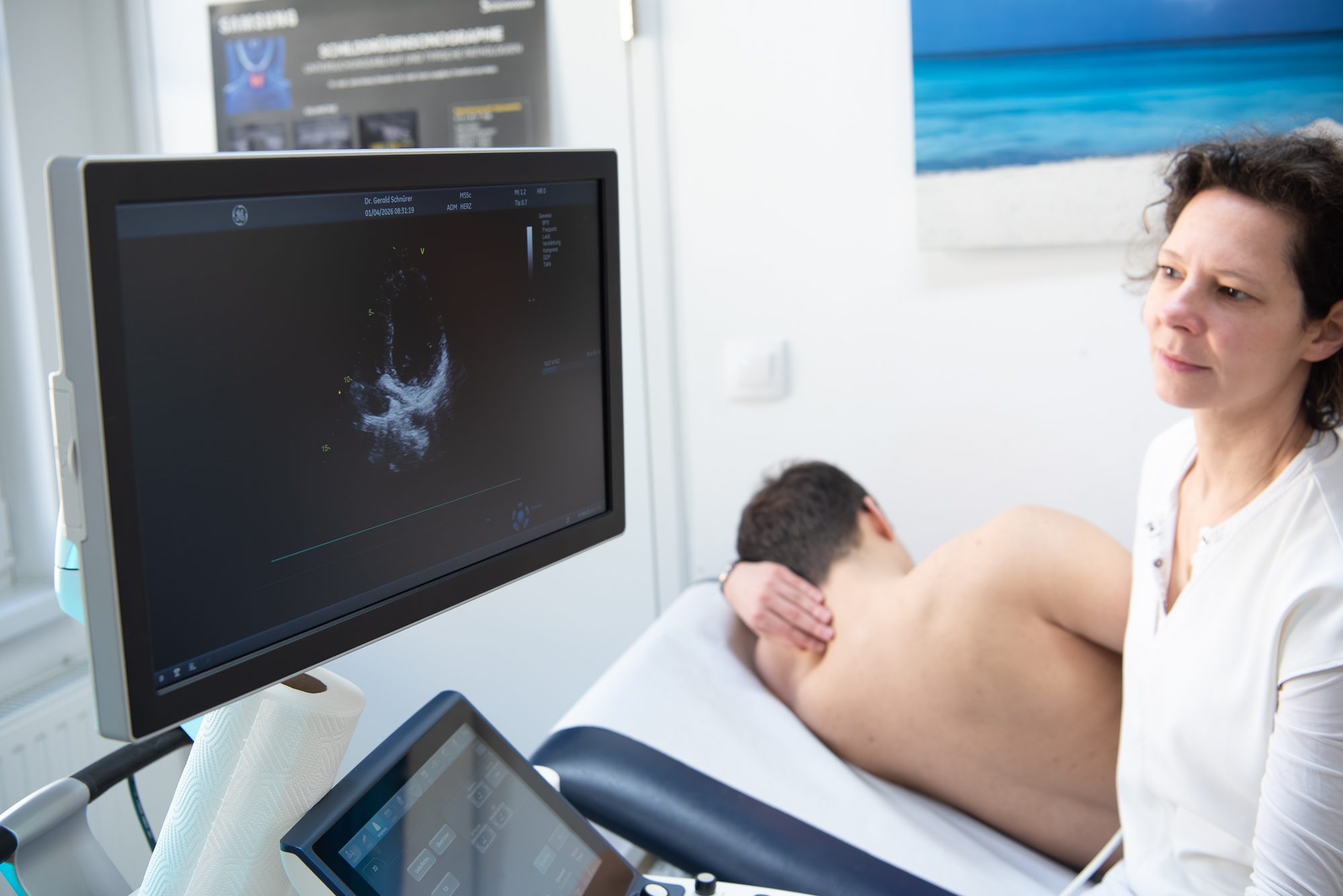 Dr. Schieder führt eine Herzultraschall-Untersuchung durch und betrachtet das Ultraschallbild am Bildschirm. Der Patient liegt seitlich, den Kopf abgewandt.